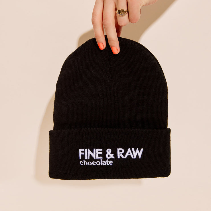 Person holding a black beanie with white FINE & RAW chocolate logo on it