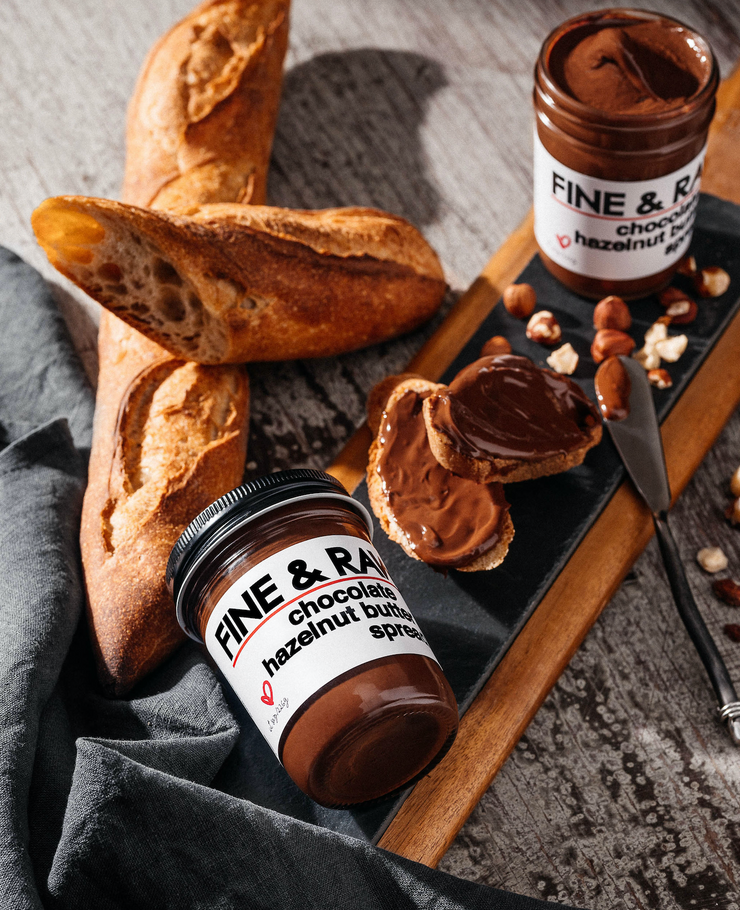 Jar of Chocolate Hazelnut Butter Spread closed and one opened. Jars, toast with spread and hazelnuts on top of a table. A knife and baguettes surround table